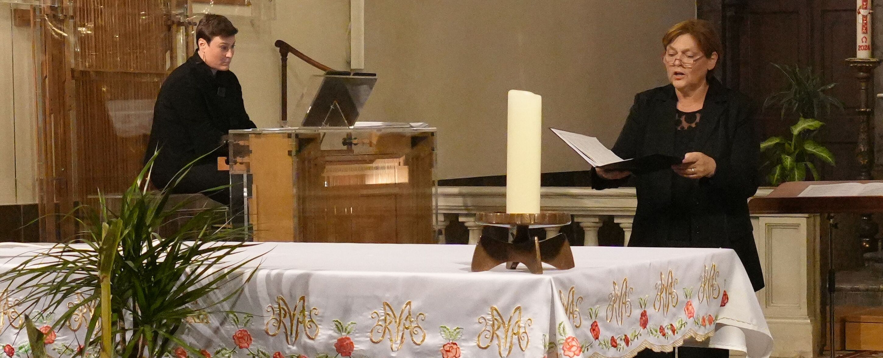 Zwei Frauen musizieren in einer Kirche; die eine spielt Orgel, die andere singt aus einem Buch.