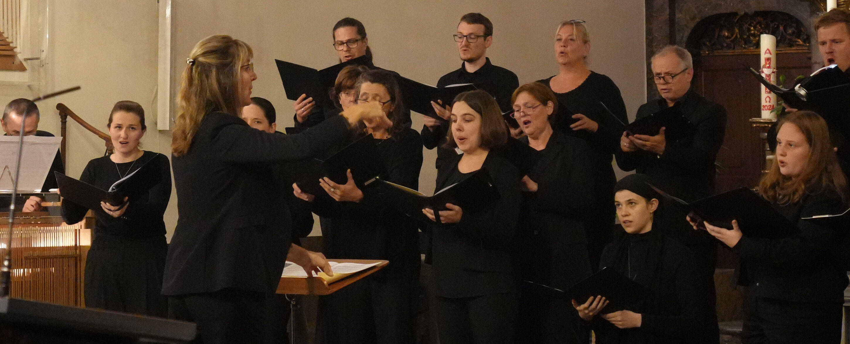 In einer Kirche tritt ein Chor unter der Leitung eines Dirigenten auf, alle in schwarzer Kleidung.