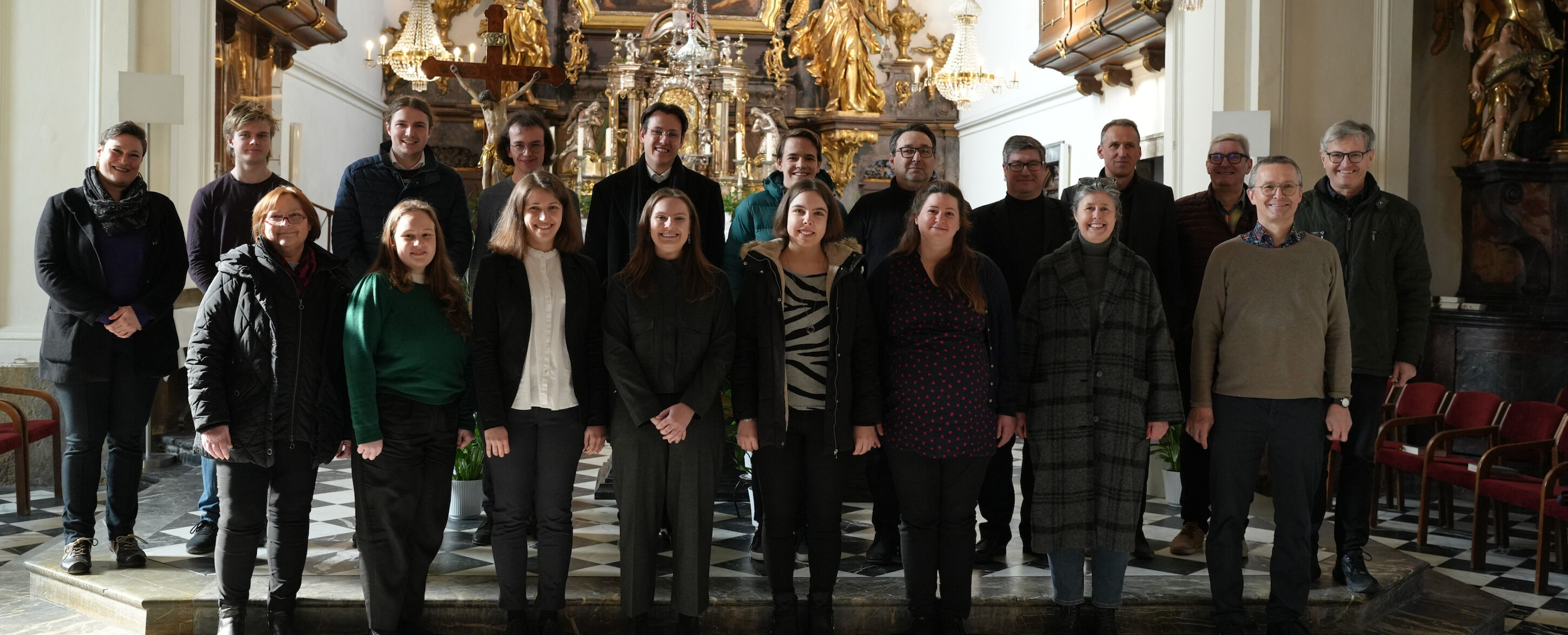 Eine Gruppe von Menschen steht zusammen in einer Kirche mit kunstvollen Verzierungen im Hintergrund.
