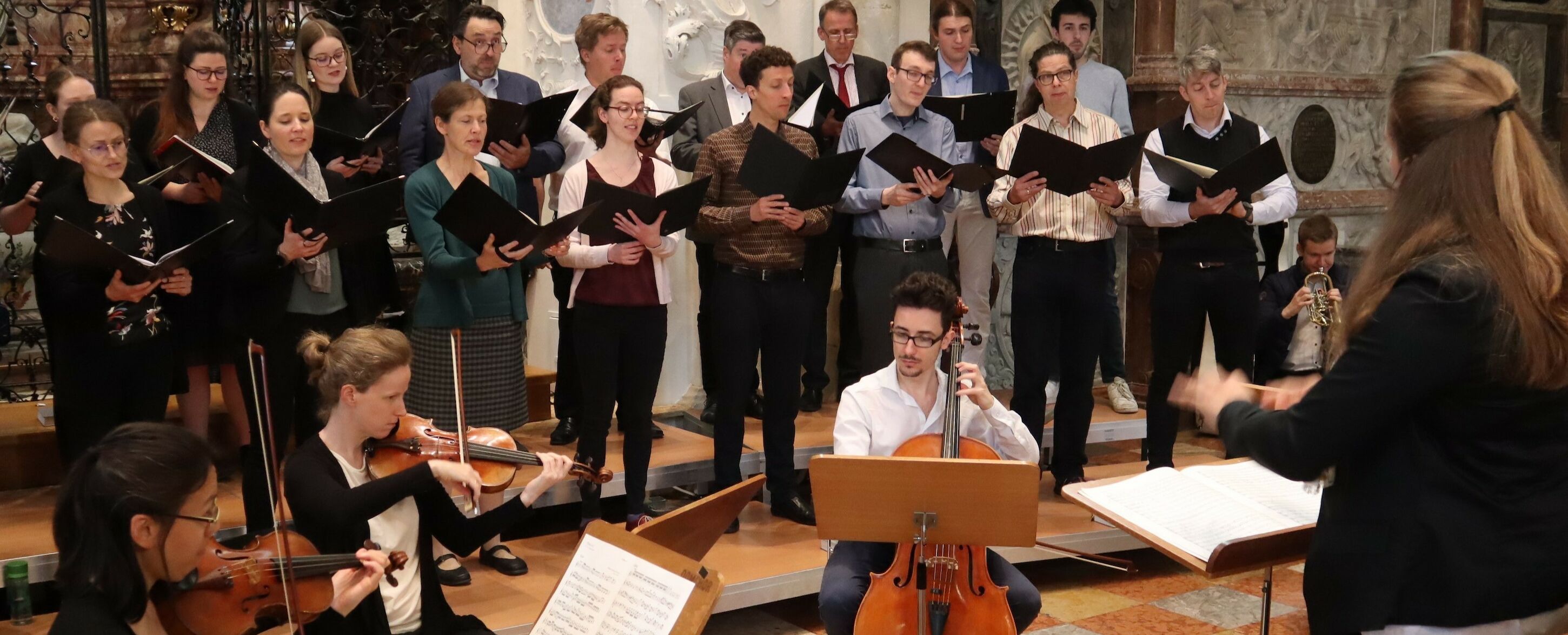 Ein Chor und Streicher treten gemeinsam mit einem Dirigenten in einem Innenraum auf.