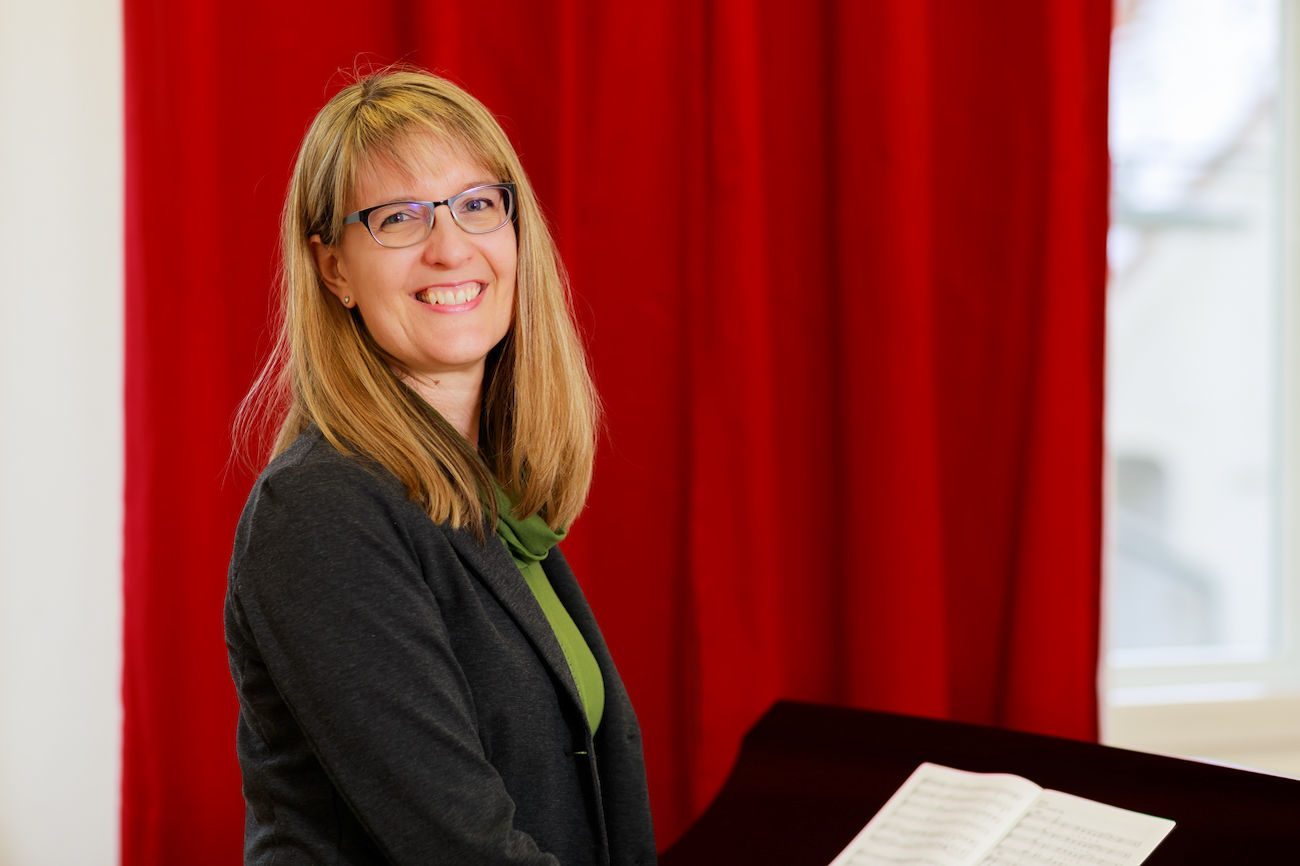 Lächelnde Frau mit Brille steht neben einem Klavier und Notenblättern, im Hintergrund ein roter Vorhang.