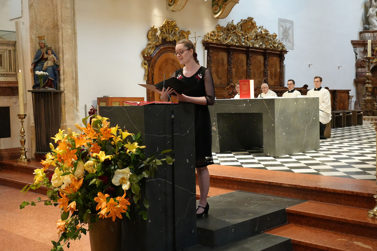 Frau im schwarzen Kleid liest an einem Kirchenpult, hinter ihr sitzen Geistliche am Altar.