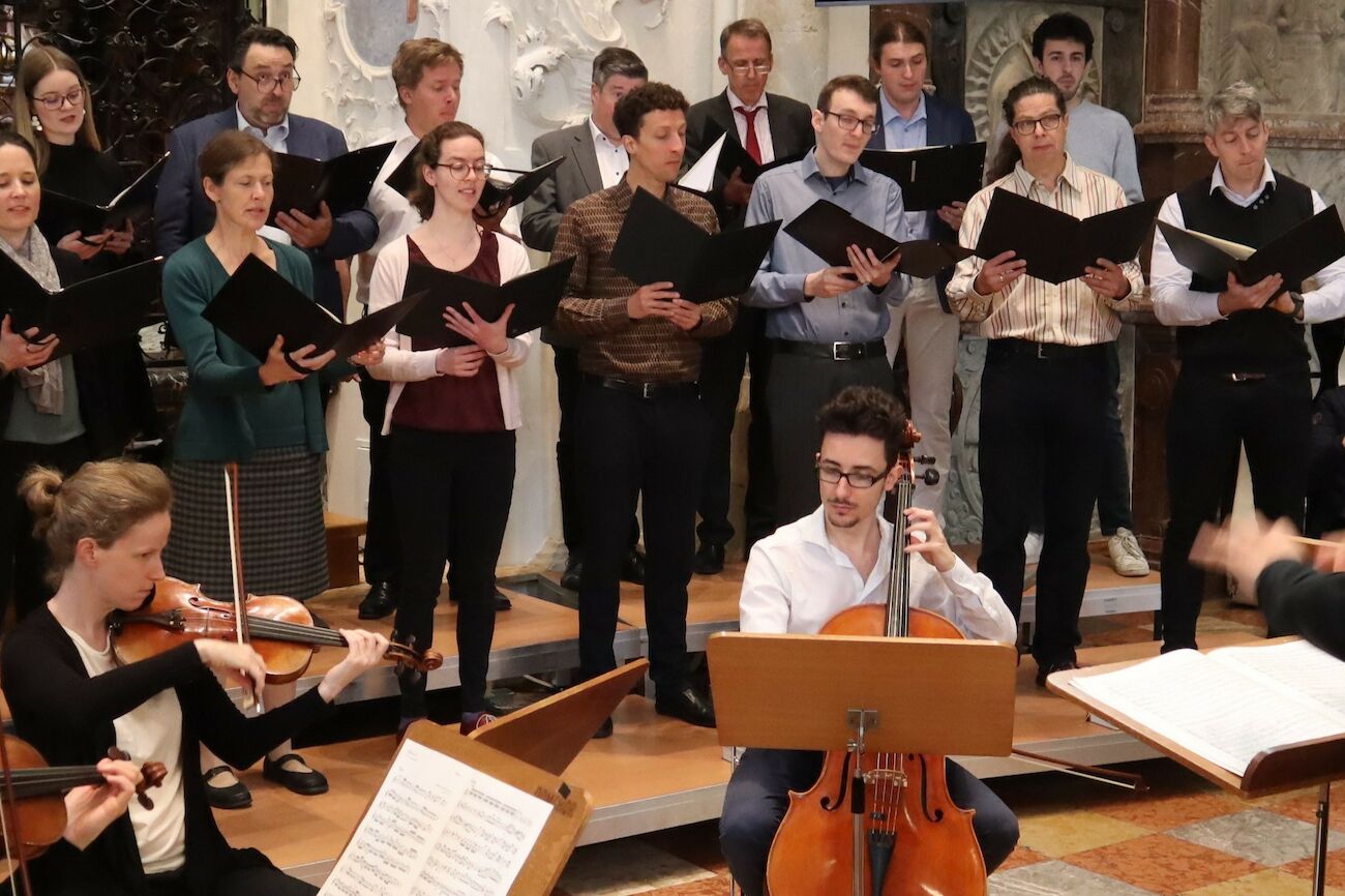 Ein Chor und Streichmusiker treten gemeinsam auf einer Bühne in einem formellen Veranstaltungsort auf.